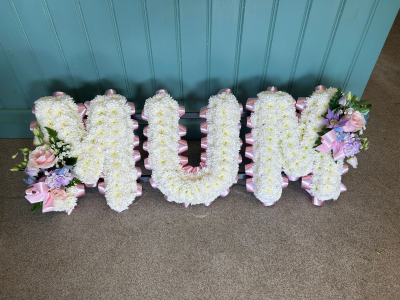Letters - A heartfelt floral letter tribute created to honour the memory of a much-loved mother with elegance and love. Finished with soft pastel accents, delicate ribbon edging, and complementary floral sprays, this touching design offers a timeless expression of remembrance. Thoughtfully handcrafted by our skilled florists using the freshest seasonal blooms and foliage, each tribute is made with great care and compassion, providing a meaningful centrepiece for funeral services, memorials, or celebration of life ceremonies.