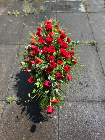 Red Rose Spray - A striking and elegant casket spray designed of Red Roses, creating a powerful tribute to a life deeply loved and remembered. Arranged in a classic teardrop shape with flowing greenery, this handcrafted design conveys love, respect, and everlasting devotion. Its timeless style makes it a meaningful choice for funeral services or celebrations of life, offering comfort and a heartfelt expression of sympathy. Each tribute is individually created by our florists using the freshest seasonal materials, ensuring every arrangement is unique while maintaining its graceful and dignified appearance.